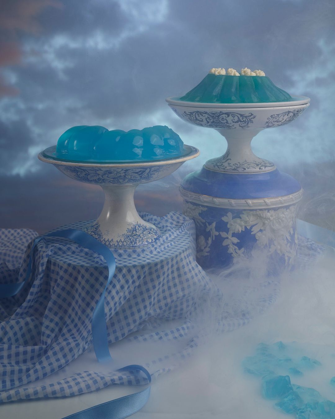 Still life food photography featuring two blue jello desserts on pedestal serving dishes, with a blue ribbon and gingham cloth.