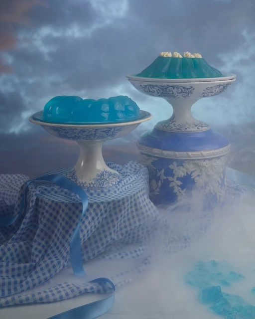 Still life food photography featuring two blue jello desserts on pedestal serving dishes, with a blue ribbon and gingham cloth.