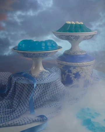 Still life food photography featuring two blue jello desserts on pedestal serving dishes, with a blue ribbon and gingham cloth.
