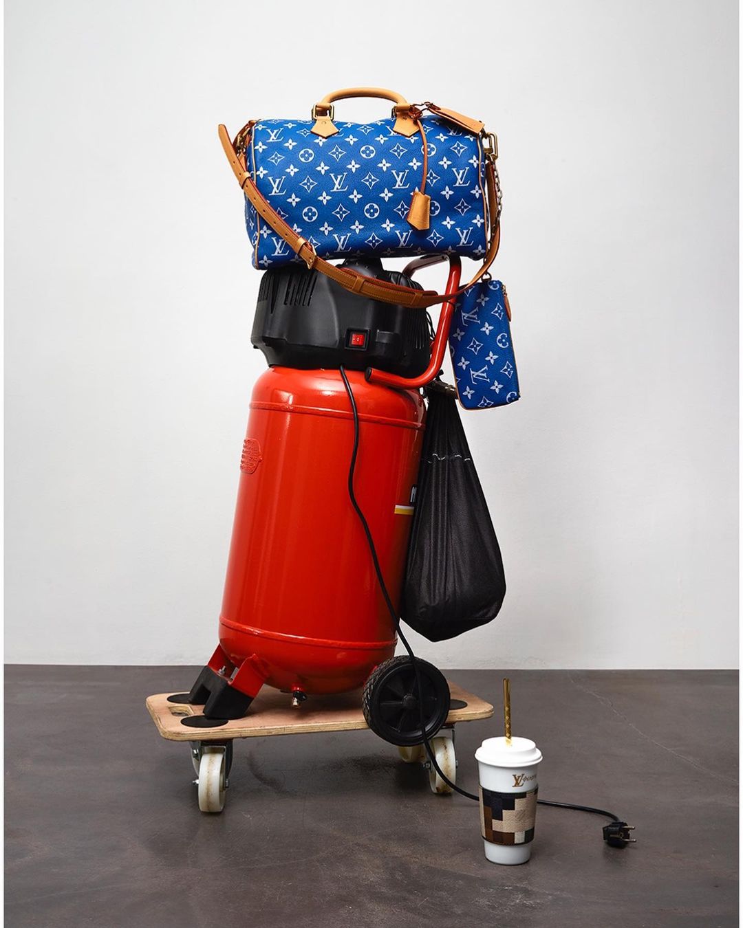 A blue and white Louis Vuitton purse stacked on top of a red air compressor on a wood platform with wheels, next to a coffee cup.