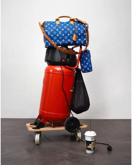 A blue and white Louis Vuitton purse stacked on top of a red air compressor on a wood platform with wheels, next to a coffee cup.