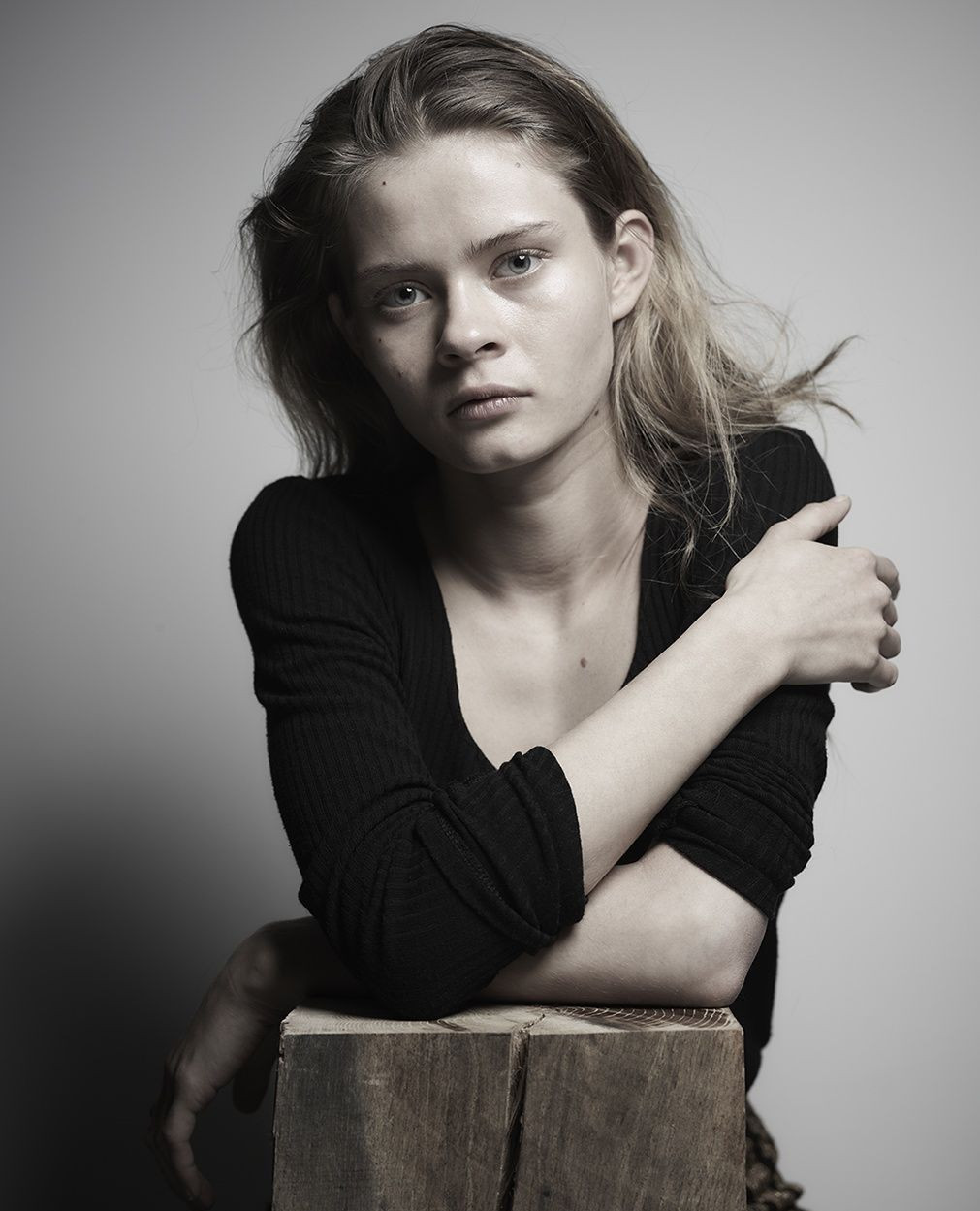 Indoor portrait of a young fair-skinned woman with blonde hair wearing a black top, posing with her arms crossed on a wood block.