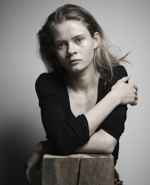 Indoor portrait of a young fair-skinned woman with blonde hair wearing a black top, posing with her arms crossed on a wood block.