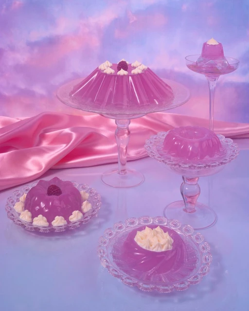 Several decorative gelatin desserts on clear glass stands, with a pink satin drape and a dreamy sky background, food photography.