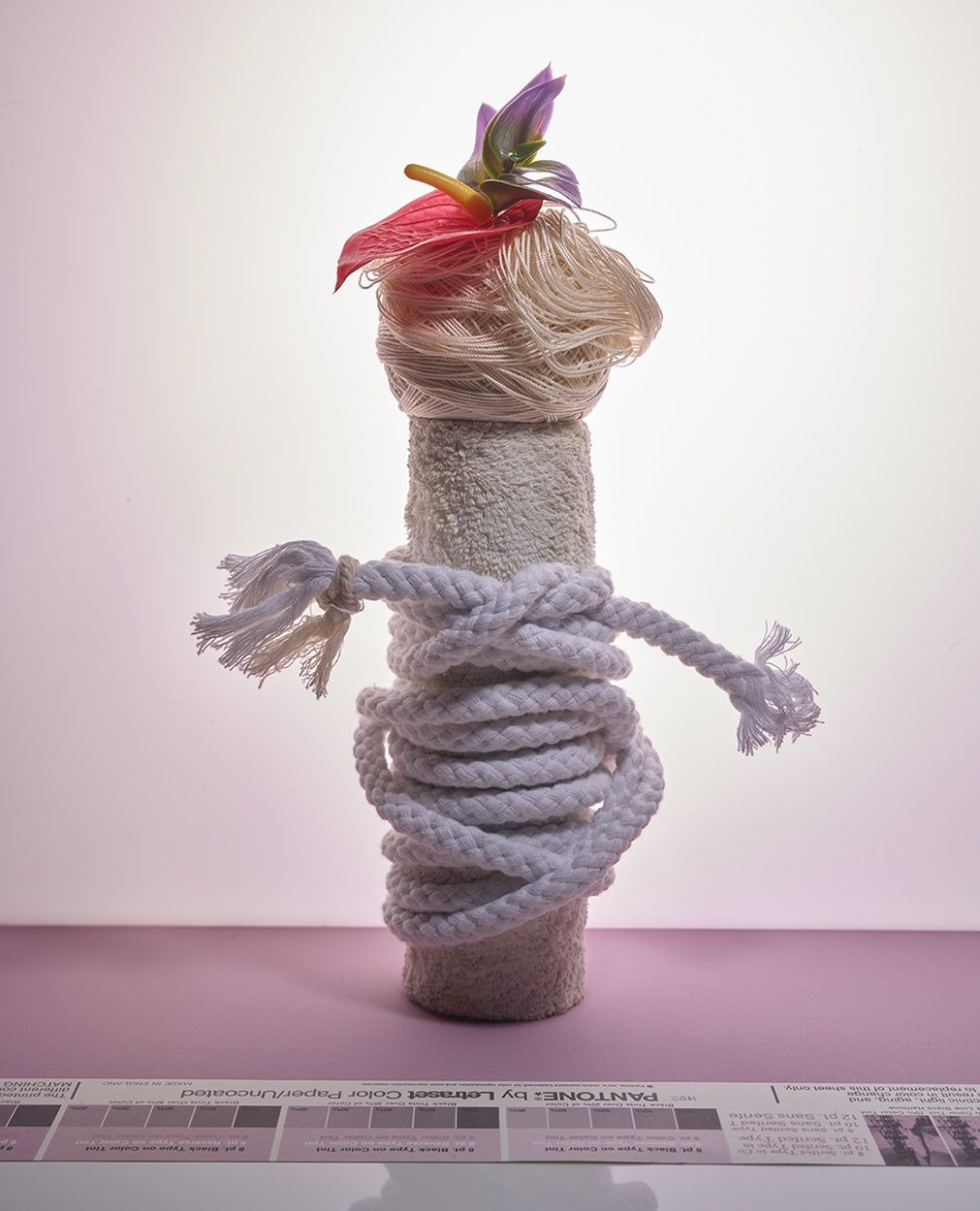 Artistic still life featuring a vertically stacked arrangement of a towel roll, wrapped rope, yarn, and flowers on a pink surface.