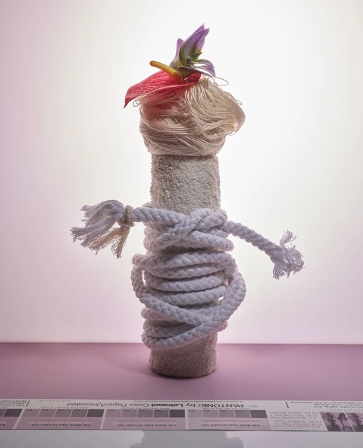 Artistic still life featuring a vertically stacked arrangement of a towel roll, wrapped rope, yarn, and flowers on a pink surface.