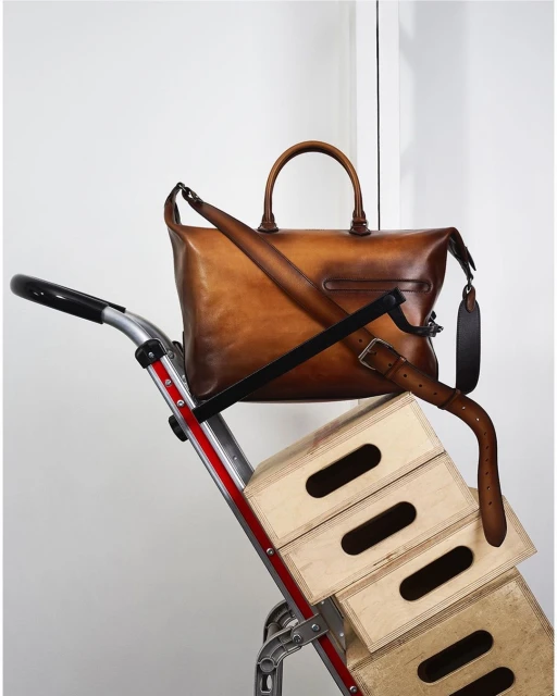 Leather duffel bag atop stacked wooden crates on a silver hand truck, indoors, against a white wall.