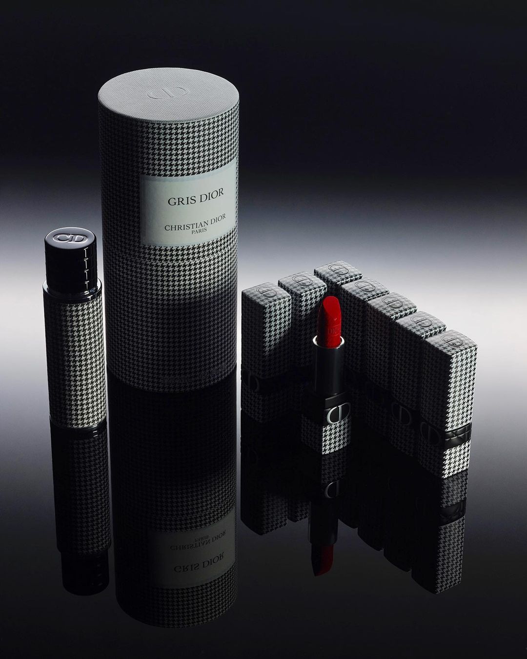 Close-up studio shot of Dior makeup and fragrances. The products feature black and white houndstooth patterns and reflect on a shiny surface.