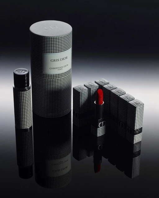 Close-up studio shot of Dior makeup and fragrances. The products feature black and white houndstooth patterns and reflect on a shiny surface.