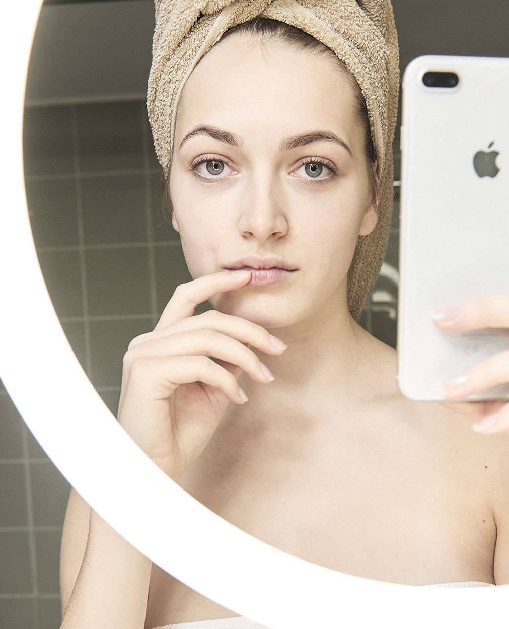 A woman in a towel poses in front of a mirror, taking a selfie with an iPhone in a bathroom with grey tiled walls.