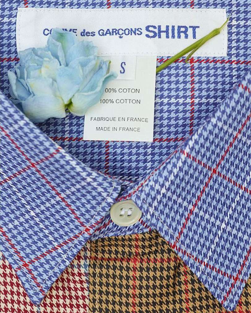 Close-up of a COMME des GARÇONS SHIRT label and garment featuring a blue and brown plaid pattern, with a light blue flower accent.