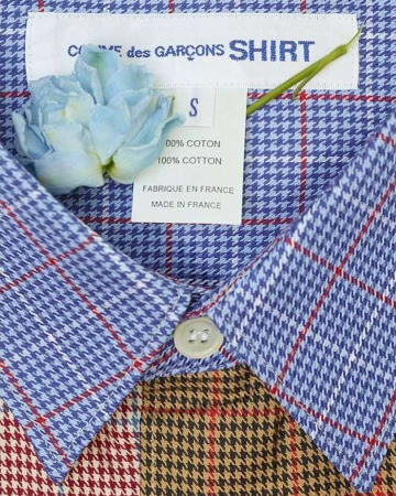 Close-up of a COMME des GARÇONS SHIRT label and garment featuring a blue and brown plaid pattern, with a light blue flower accent.