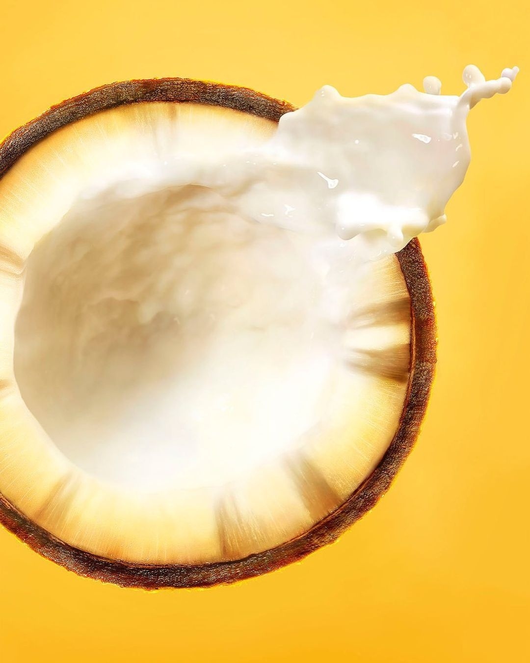 A halved coconut with white liquid splashing out against a solid yellow background, showcasing fresh tropical fruit and vibrant color.