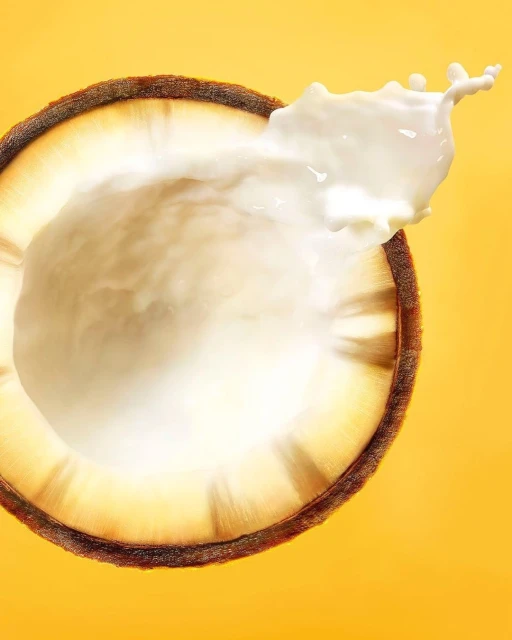 A halved coconut with white liquid splashing out against a solid yellow background, showcasing fresh tropical fruit and vibrant color.