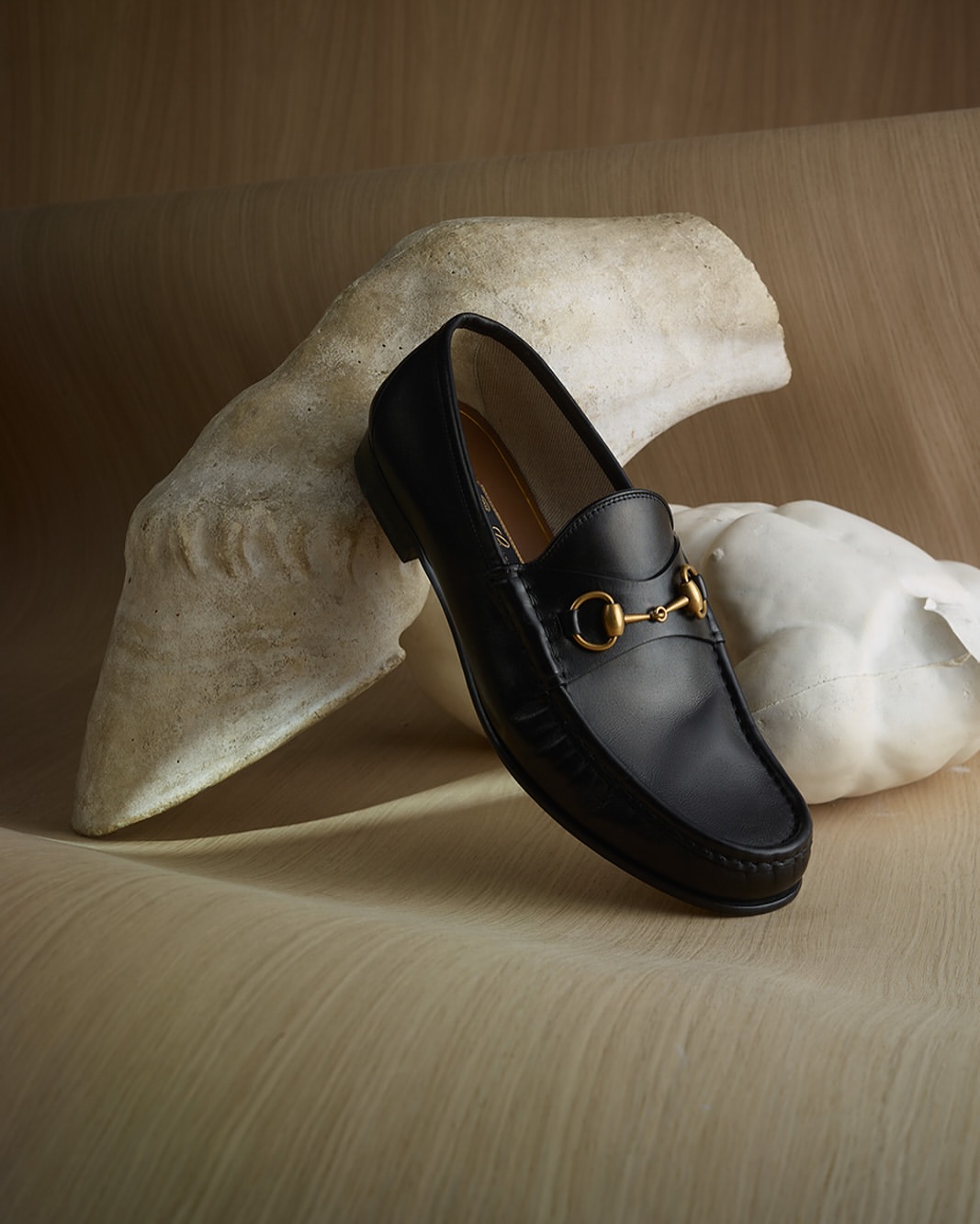 Elegant black leather Gucci loafer with gold horse-bit detail next to a light-colored, textured backdrop and sculptural form.