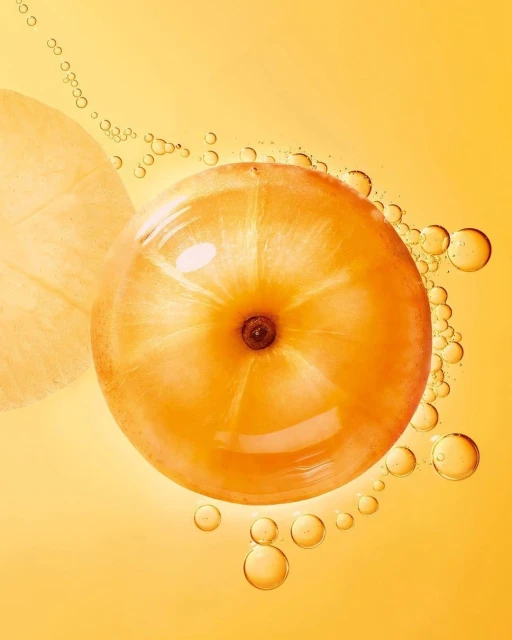 Close-up of a whole amla fruit surrounded by oil droplets on a gradient yellow background, suitable for beauty or food-related themes.