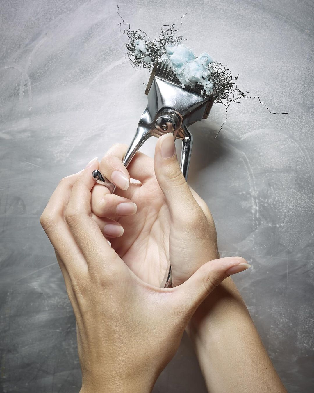 Close-up of hands holding a vintage metal hair clipper with hair and cotton caught in the blades, set against a textured gray backdrop.