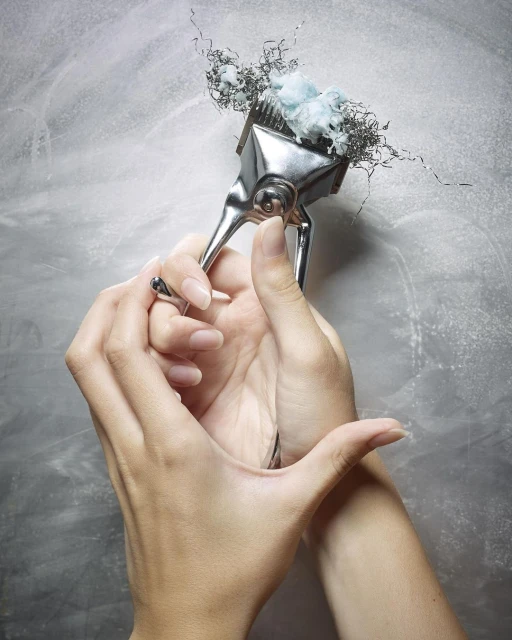 Close-up of hands holding a vintage metal hair clipper with hair and cotton caught in the blades, set against a textured gray backdrop.