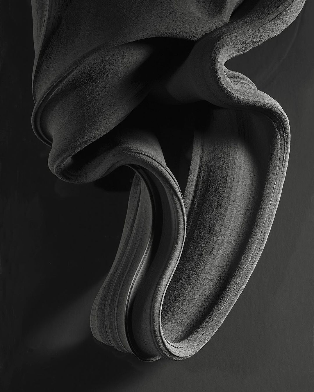 Abstract gray fabric sculpture with textured surface and soft lighting on a dark background, creating depth and movement.