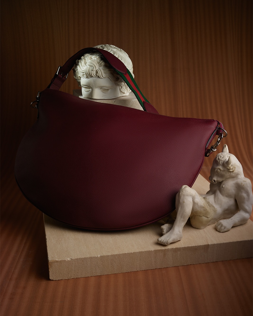 Burgundy leather hobo bag with a striped strap rests on a light brown wood-like pedestal next to two sculpture busts.