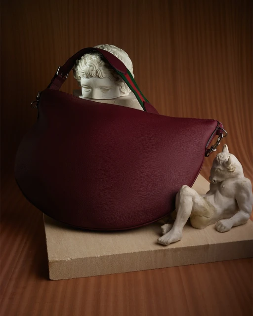Burgundy leather hobo bag with a striped strap rests on a light brown wood-like pedestal next to two sculpture busts.