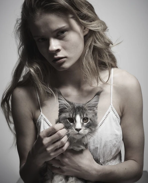 Monochrome studio portrait of a young adult female holding a fluffy grey tabby cat in her arms with a neutral facial expression.