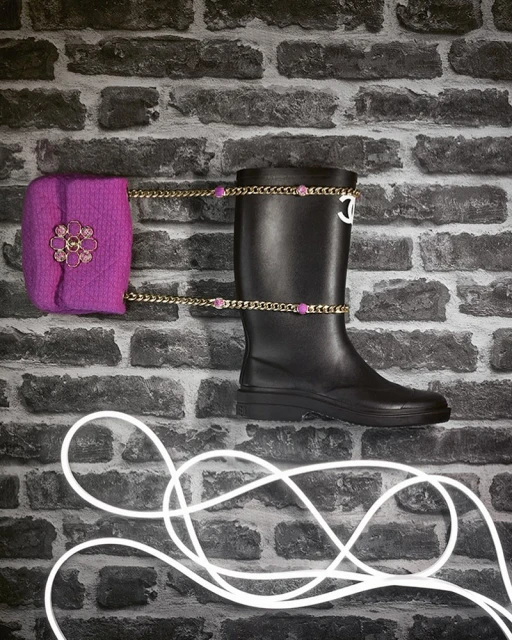 A black rubber boot and a magenta purse are connected by a gold chain, set against a grey brick wall and illuminated by a glowing white wire.