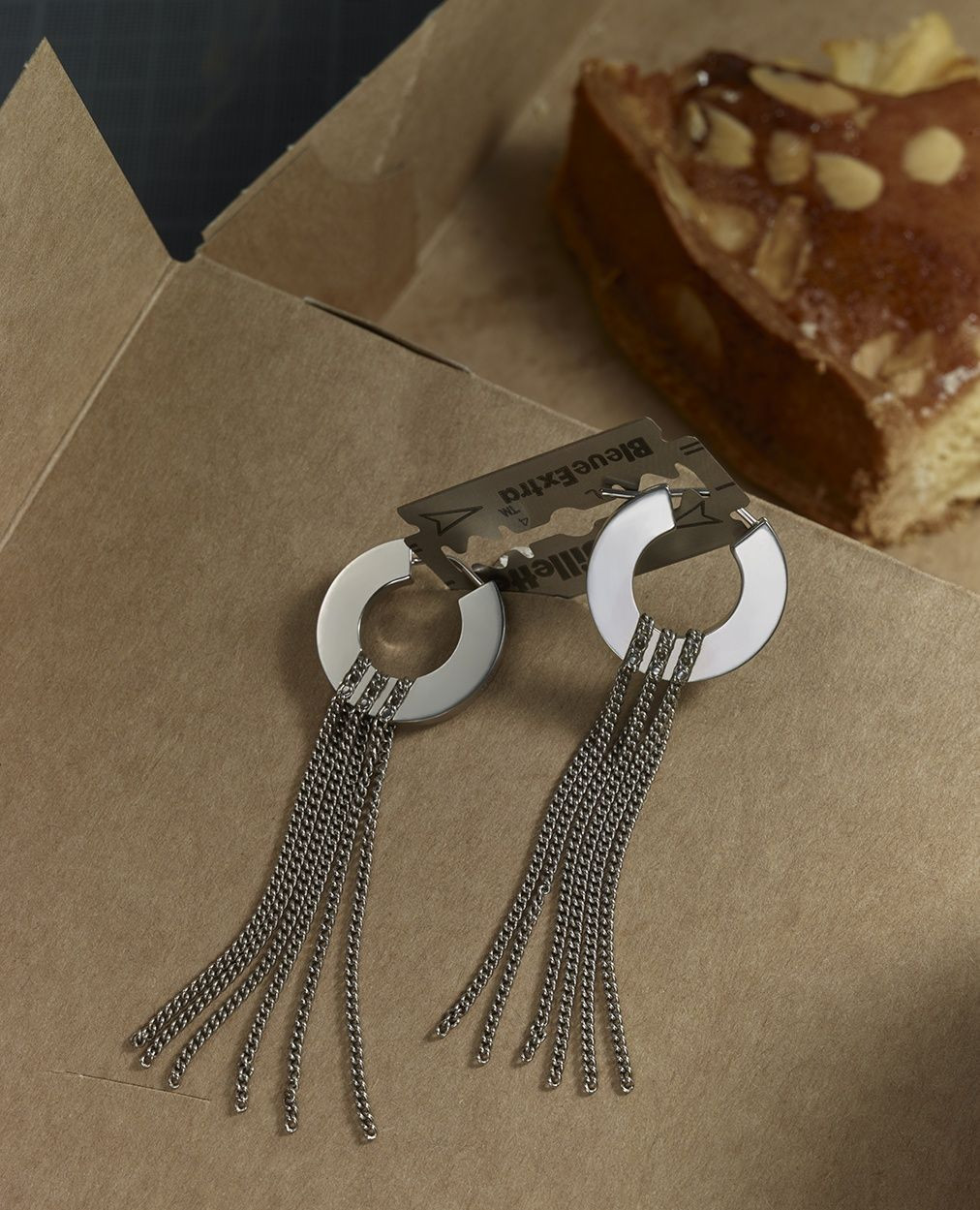 Stylish silver-tone earrings with chain fringe and a cake slice and razor blade on a cardboard surface, adding an edgy touch.