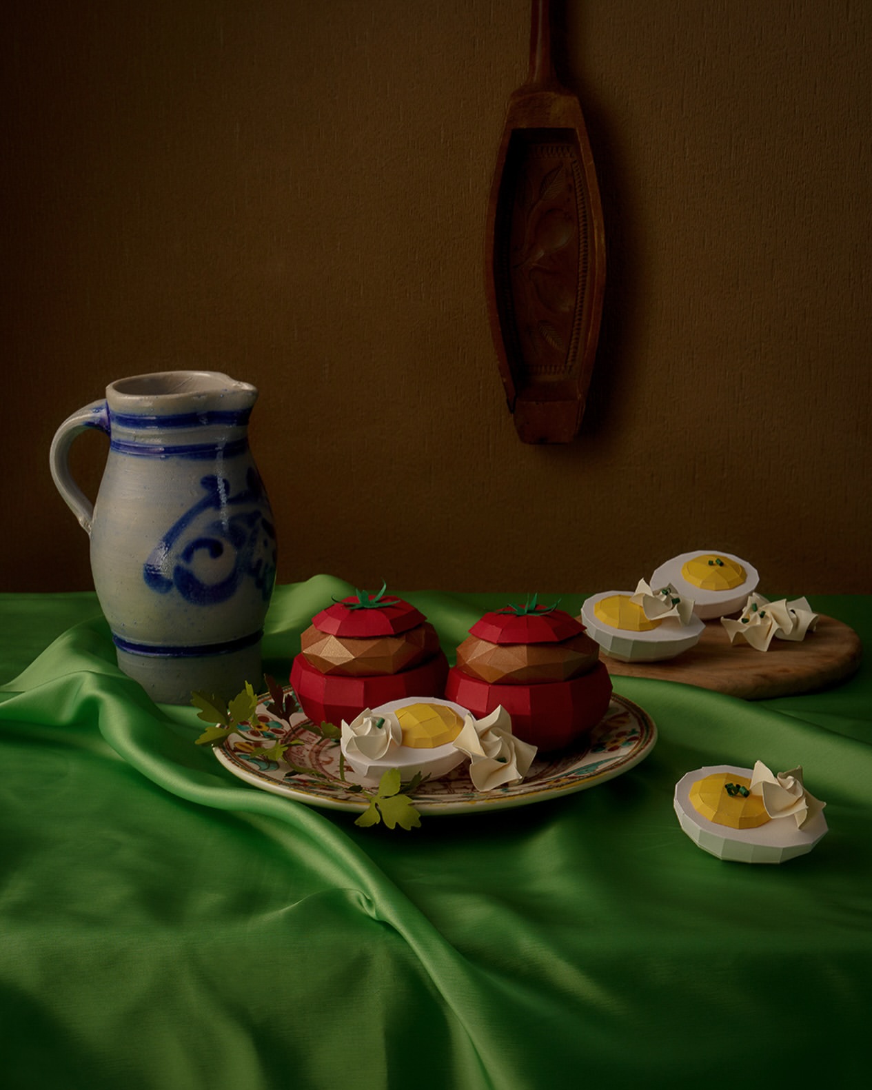 Still life of two tomato-shaped containers and egg halves on a green silk tablecloth, featuring a pitcher and wooden spoon in the background.