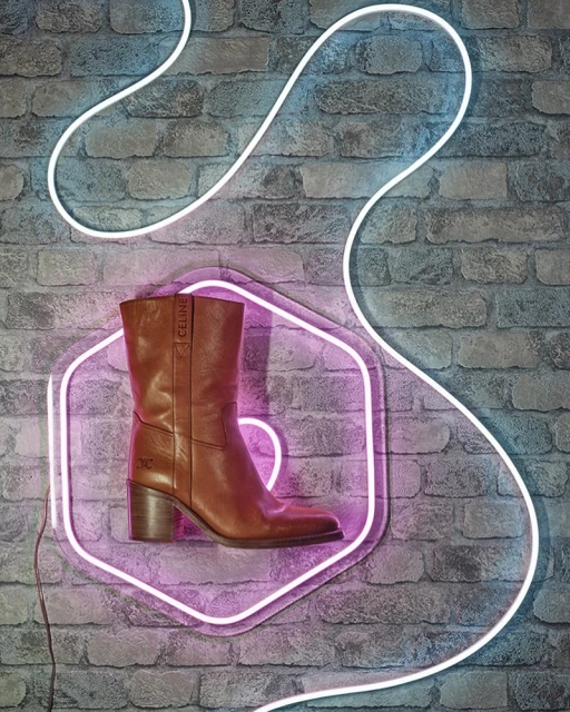 Leather boot by Celine stands on a neon-lit platform against a brick wall, combining fashion and modern lighting.