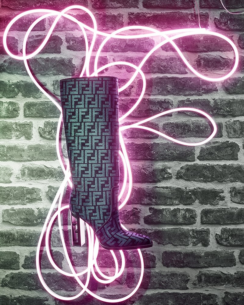 High-heeled boot with Fendi pattern displayed against a brick wall and neon pink lights, creating a high-fashion studio shot.