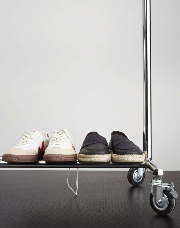 Two pairs of shoes, including sneakers and loafers, are arranged on a metal rack with wheels against a plain, bright studio backdrop.