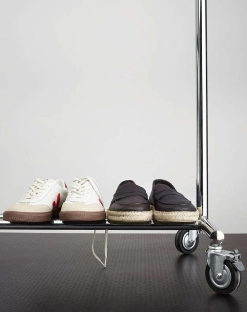 Two pairs of shoes, including sneakers and loafers, are arranged on a metal rack with wheels against a plain, bright studio backdrop.
