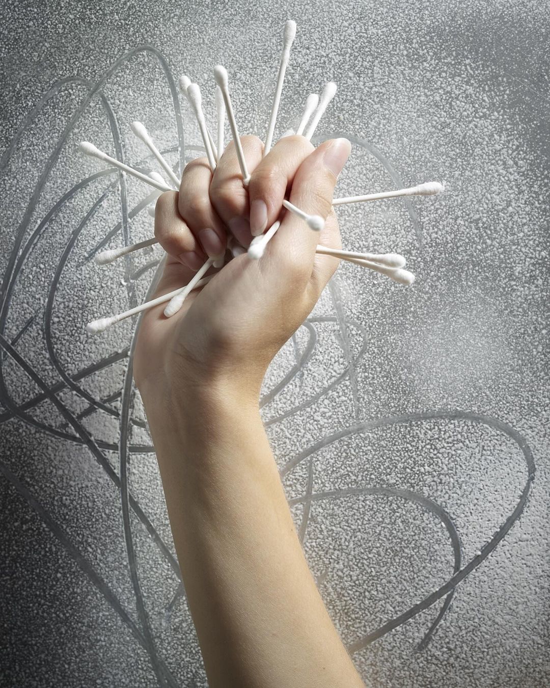 Hand holding multiple cotton swabs against a silver background. Conceptual healthcare or hygiene image with stark lighting and texture.