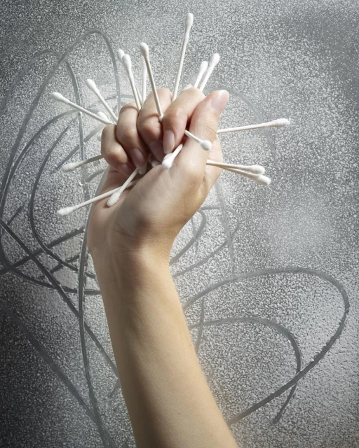 Hand holding multiple cotton swabs against a silver background. Conceptual healthcare or hygiene image with stark lighting and texture.