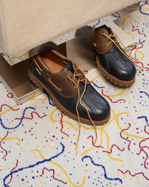 A pair of Sperry duck boots with dark blue rubber soles and brown leather uppers sit on a colorful, patterned rug next to a table.