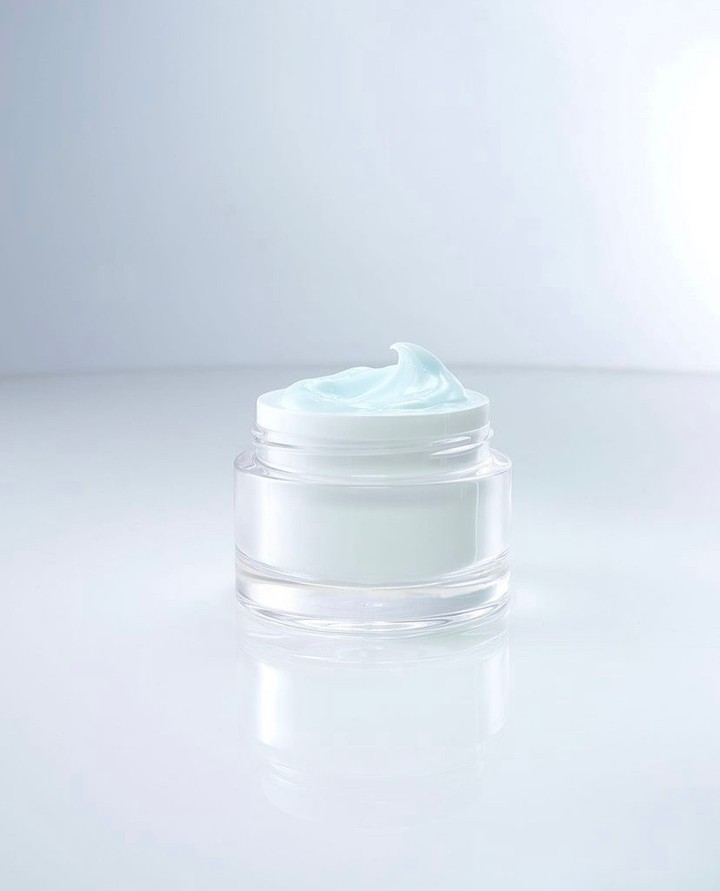 A clear jar of moisturizer sits on a reflective surface against a gradient white background, showcasing its smooth texture and refreshing color.