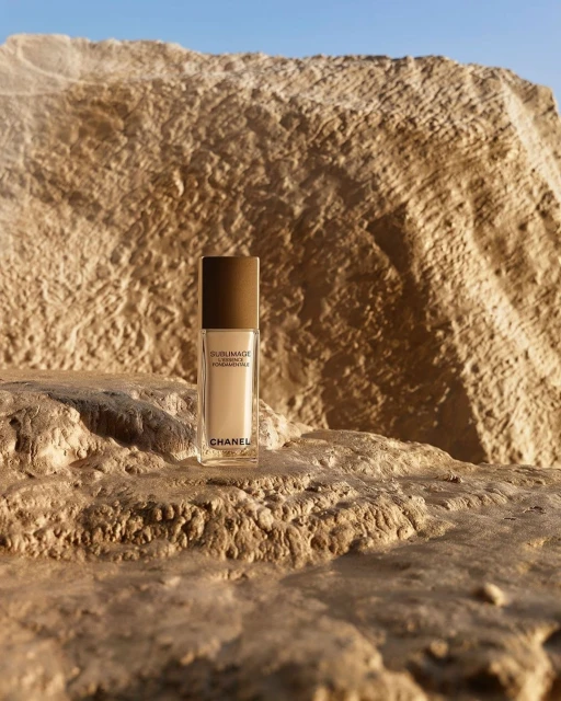 Chanel foundation bottle stands on a textured rock surface, set against a sunlit stone formation under a clear blue sky. Beauty product shot.