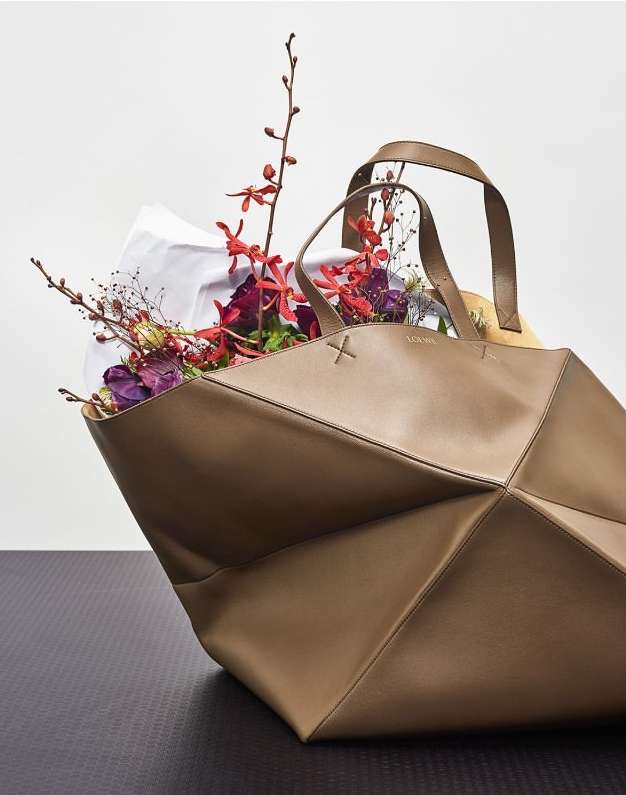 Geometric leather tote bag in a neutral tone, filled with colorful flowers and a white liner, resting on a dark, textured surface.