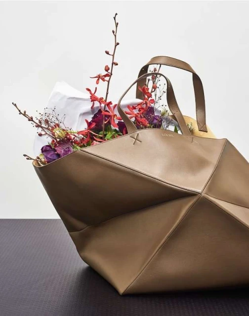 Geometric leather tote bag in a neutral tone, filled with colorful flowers and a white liner, resting on a dark, textured surface.