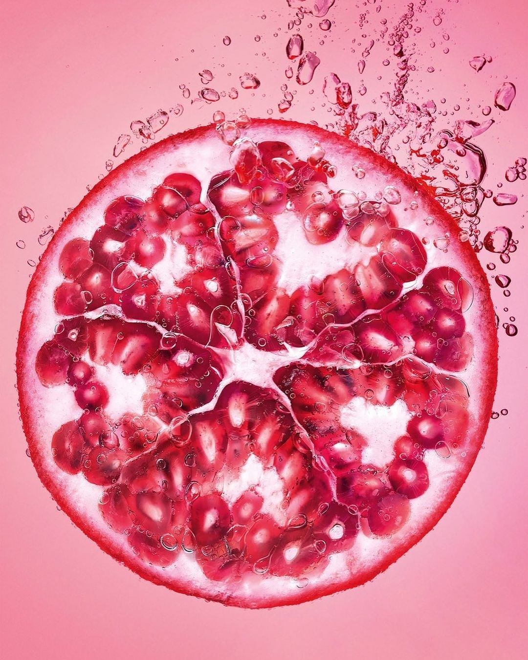 Macro shot of a vibrant pomegranate half, its ruby-red seeds glistening, against a soft pink backdrop, highlighting its juicy texture.
