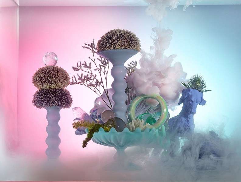 Studio shot of a surreal sculpture featuring sea urchins, glass ornaments, a blue dog figurine, and smoke effects in a pink and blue gradient.