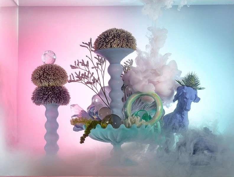 Studio shot of a surreal sculpture featuring sea urchins, glass ornaments, a blue dog figurine, and smoke effects in a pink and blue gradient.
