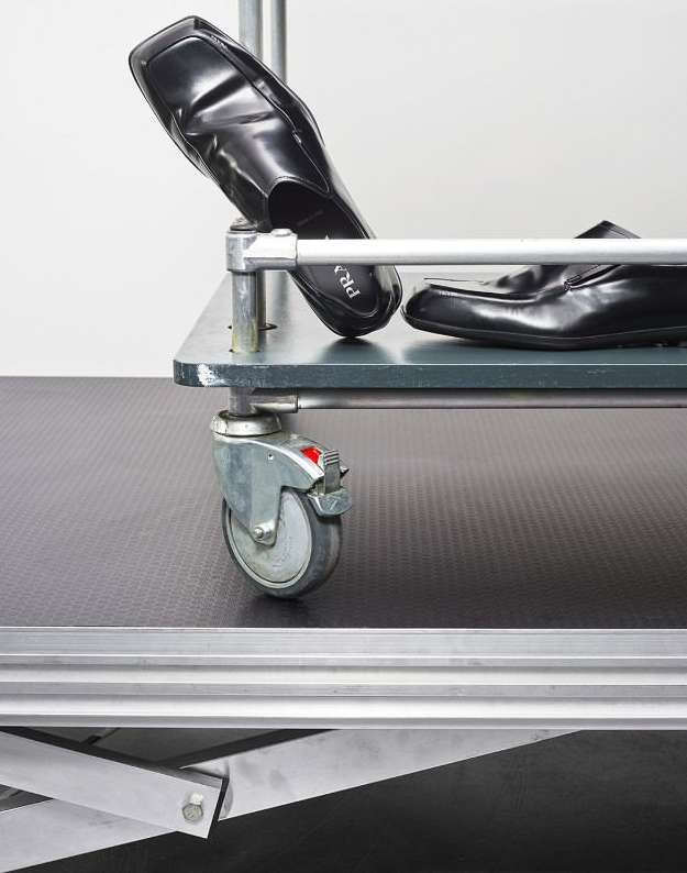 Two black leather loafers sit on a grey metal shelving unit with caster wheels against a plain white background in a studio setting.
