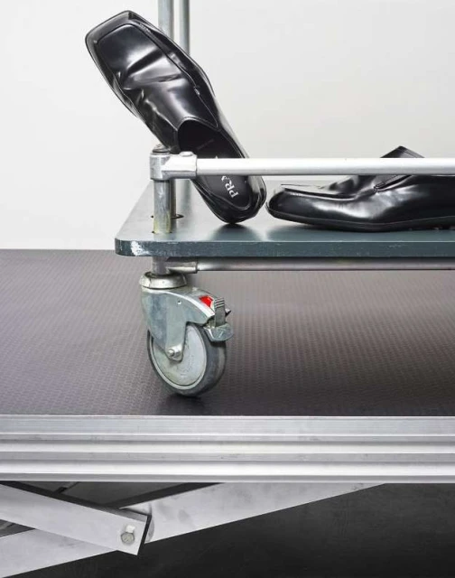Two black leather loafers sit on a grey metal shelving unit with caster wheels against a plain white background in a studio setting.