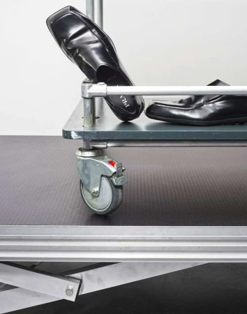 Two black leather loafers sit on a grey metal shelving unit with caster wheels against a plain white background in a studio setting.