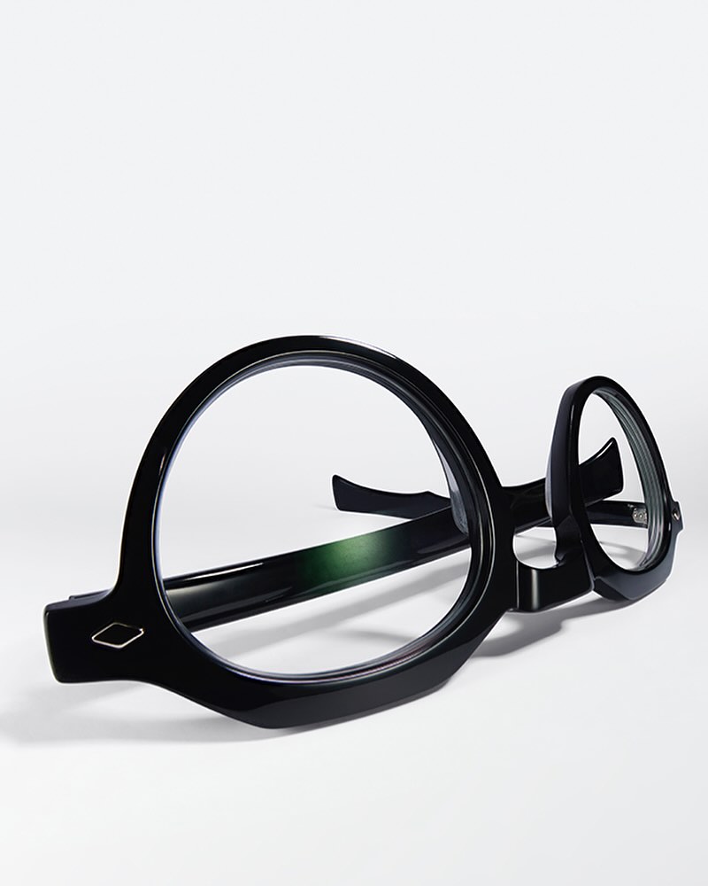 Close-up of round, glossy black eyeglasses resting on a white surface, casting a soft shadow in a brightly lit, minimalist composition.