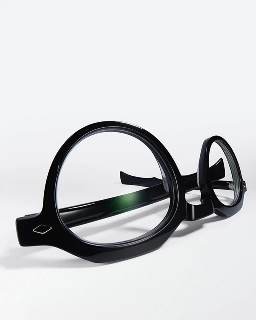 Close-up of round, glossy black eyeglasses resting on a white surface, casting a soft shadow in a brightly lit, minimalist composition.