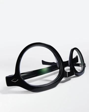 Close-up of round, glossy black eyeglasses resting on a white surface, casting a soft shadow in a brightly lit, minimalist composition.