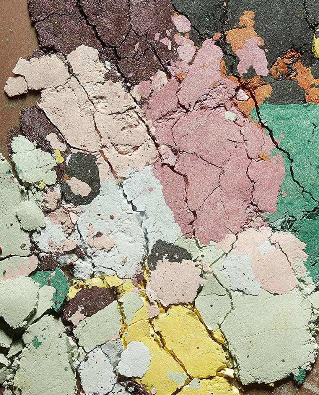 Overhead close-up captures a cracked, multicolored makeup palette in hues of green, pink, yellow, and brown with a rough texture.